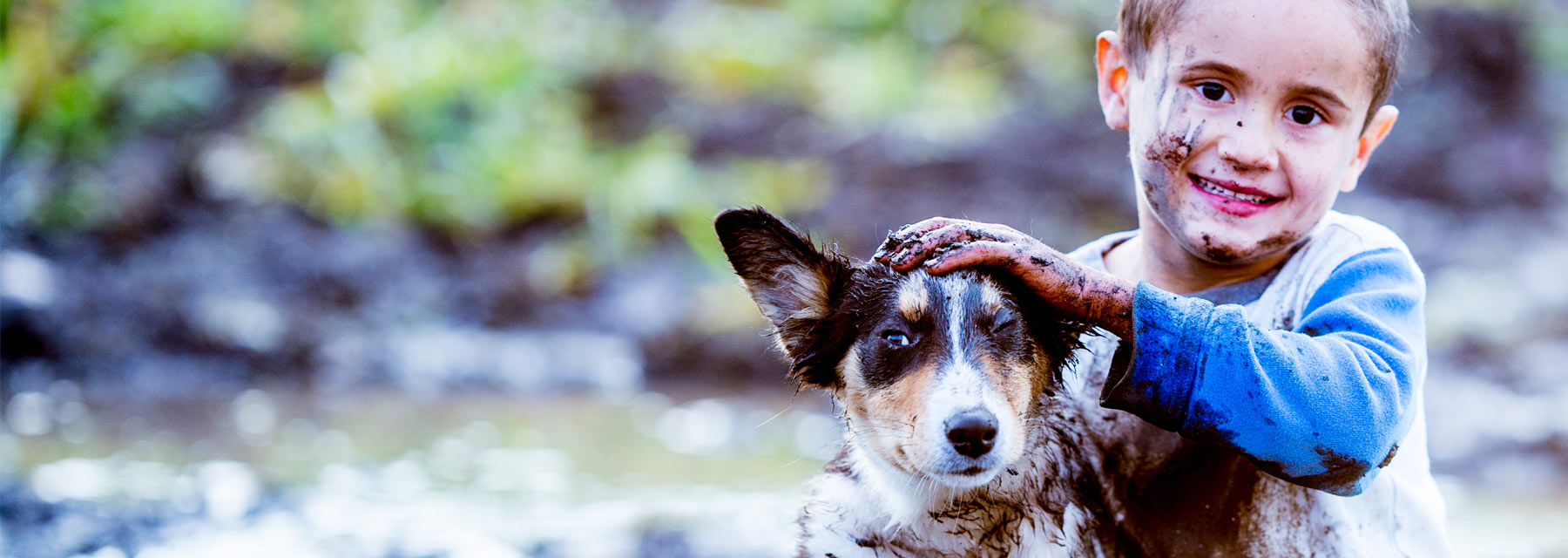 Young smiling boy wearing a muddy long-sleeved knit shirt while petting a muddy dog on a rainy day in a forest