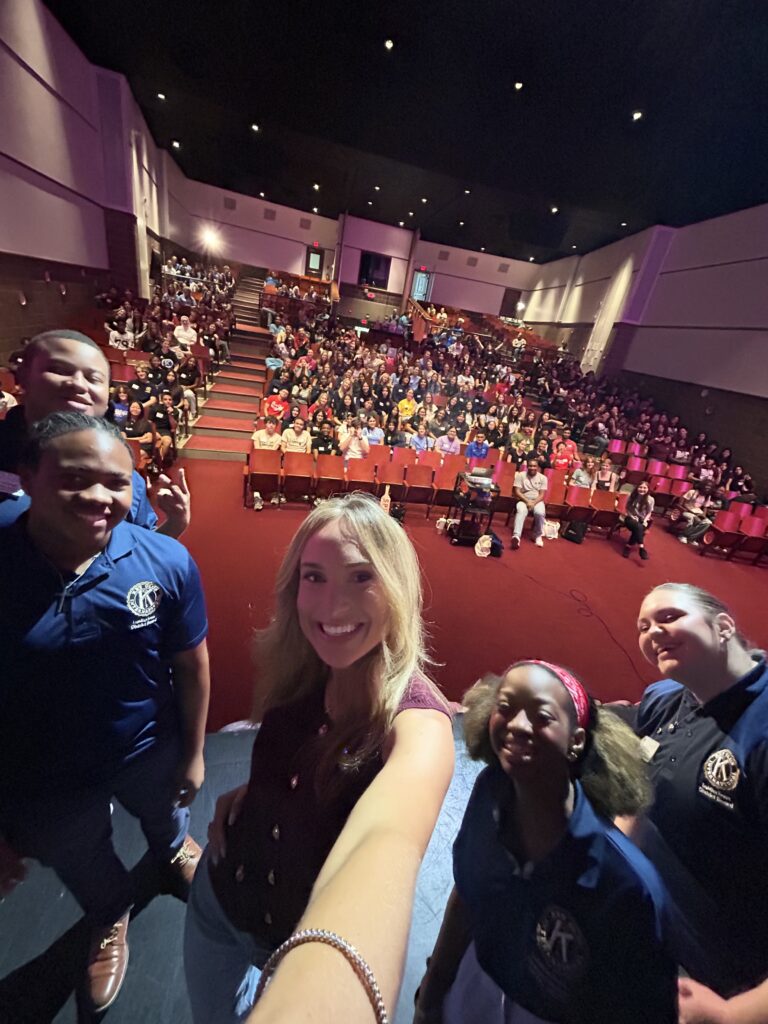 A smiling woman takes a selfie in front of a large crowded auditorium while onstage with several Key Club students