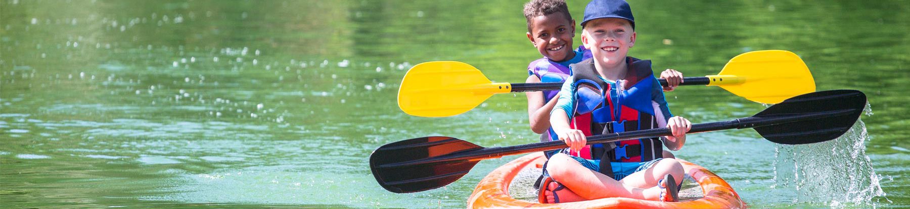 Two young smiling boys wearing multi-colored life vests while paddling in a orange kayak on a calm green lake