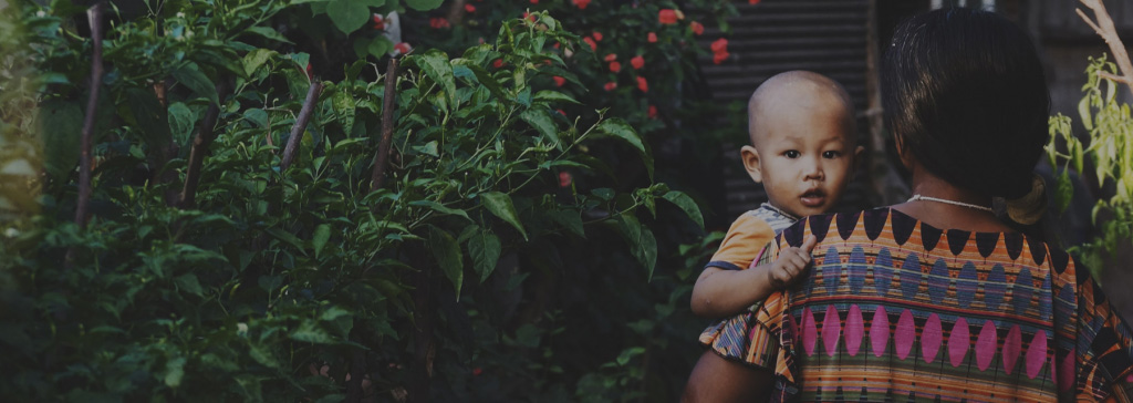 Young mother with black hair pulled in a bun, facing away, wearing a short-sleeved dress while carrying an infant in a short-sleeved shirt