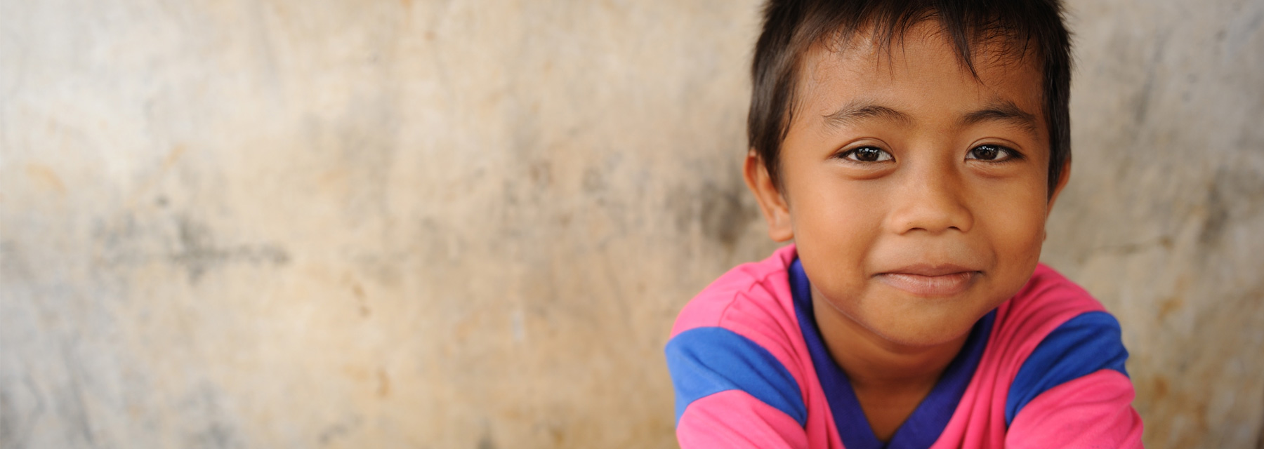 Young smiling boy with short black hair wearing a pink and blue shirt while leaning up against a smooth concrete wall