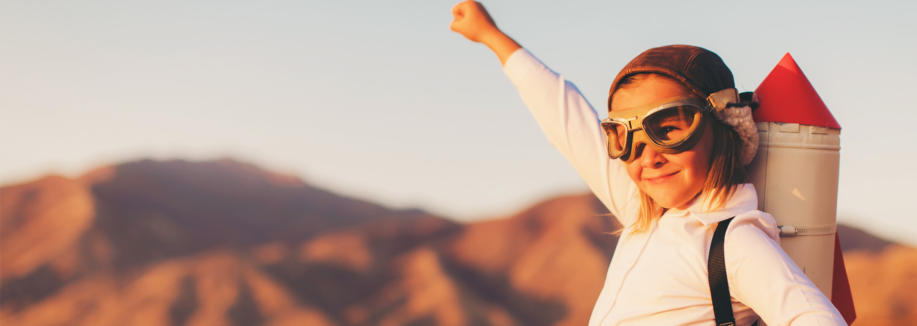 Young girl with short dark blonde hair wearing a white hoodie, brown vintage aviation googles and cap carrying a toy rocket ship on her back while raising her right hand in a victory pose