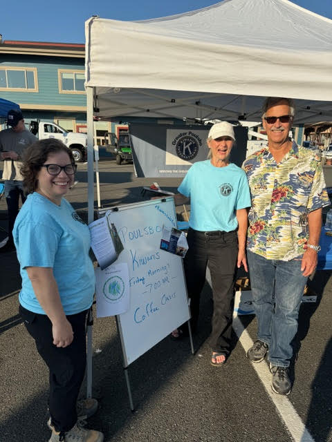 Kiwanis members are standing at their booth during National Night Out in downtown Poulsbo. They are smiling and interacting with community members, surrounded by informational materials and activities that highlight the club's commitment to service and community engagement. The booth is lively, with banners and signage promoting Kiwanis' mission.