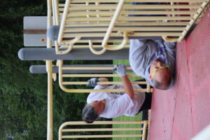 A group of Kiwanis members is seen cleaning and scrubbing playground equipment at Betty Iverson Kiwanis Park in Poulsbo, WA. The scene captures the volunteers working together to ensure the playground is clean and safe for children, with bright equipment and a backdrop of greenery visible in the park.