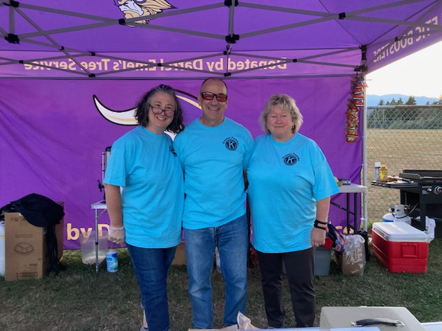 
Kiwanis members are seen volunteering at a North Kitsap High School football game, grilling hamburgers for the NKHS Booster Club. They are wearing aprons and smiles, standing by the grill as they cook and serve food, contributing to the lively and supportive atmosphere of the game.