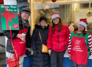 Members of Kiwanis and the NKHS Key Club are seen standing together, smiling, while ringing bells for the Salvation Army. They are dressed warmly for the winter, holding red kettles, and bringing holiday cheer as they volunteer to raise funds for the community.