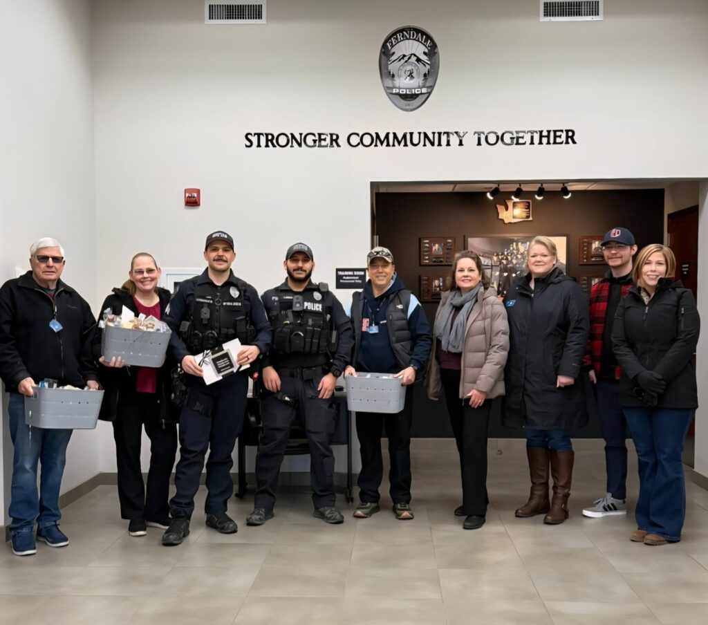 A group of 9 people standing shoulder to shoulder in the lobby of Ferndale's Police Department building under the Police Department's motto, "Stronger Community Together. Five of them are Kiwanis of Ferndal team members, two police officers, and two members of Crave Catering. From left to right, an older male Kiwanis team member is holding a basket loaded with baked items and snacks. A woman from Crave Catering holds another basket filled with the same things. Next are two male police officers, one holding a black and white See's candies box of brittle. Next is a male Kiwanis team member holding a third basket. Next are two female Kiwanis team members. Then, a young male Kiwanis team member. Last is a female member of Crave Catering.