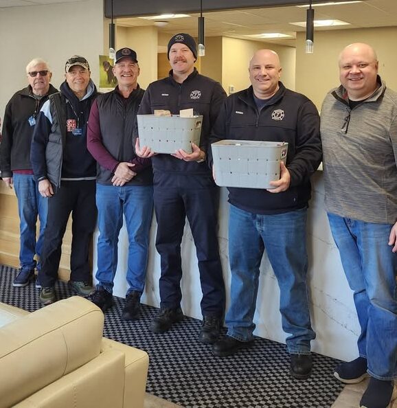 Two male Kiwanis members standing side by side with four male firefighting officials. Two of the officials are holding baskets of assorted baked goods and treats.