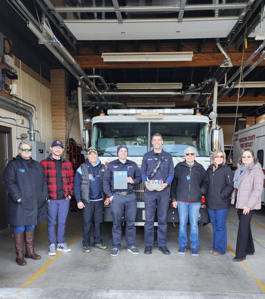 Eight people, five of whom are Kiwanis members, one with Crave Catering, and two firefighters posing in a line in front of a fire engine. One firefighter is holding a certificate of appreciation, and the other a basket of assorted baked goods and treats.