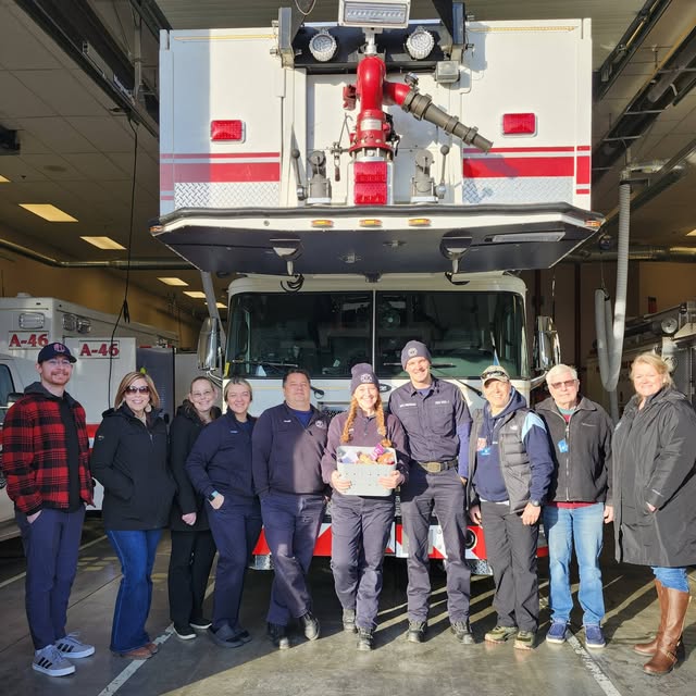 A firehouse crew and Kiwanis members wearing winter clothing are lined up in front of a fire truck. The fire woman in the center is holding a basket of assorted baked goods and treats.