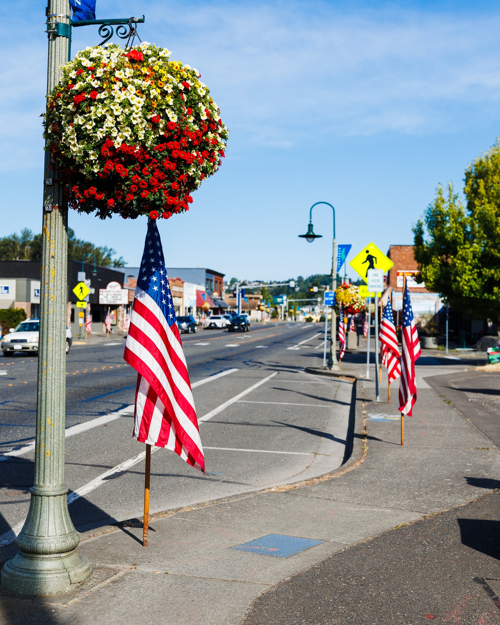 Beautiful sunny day on Main Street. The light post has a large hanging basket with green and red flowers, and American flags line Main Street's sidewalks.