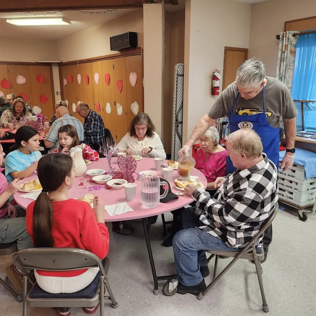 Large circular table covered in a pink tablecloth with a red heart laying in the center of the table with a double heart centerpiece on top. Seniors and fifth-grade students sit around the table sharing a turkey dinner meal while a club member pours gravy over someone's mashed potatoes.