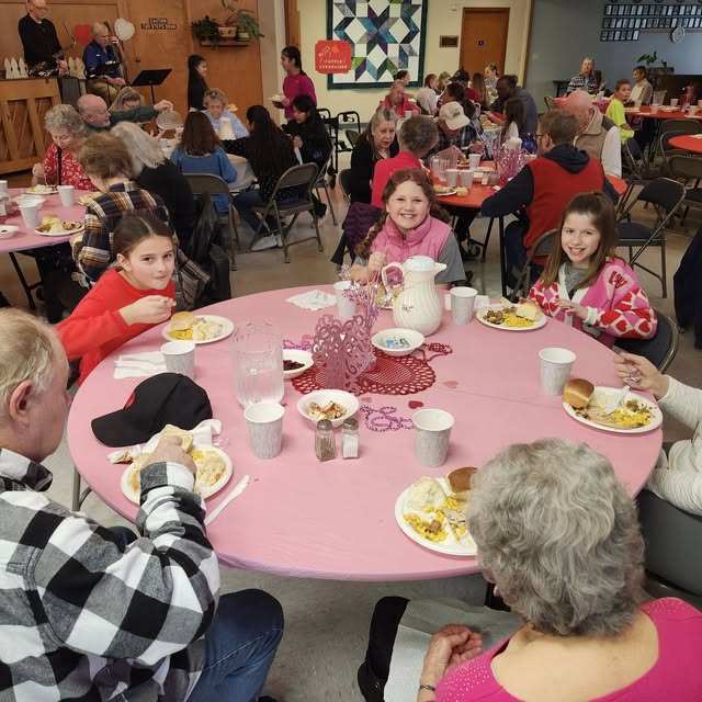 In a large lunchroom, there are six circular tables. In the foreground, three senior citizens and three fifth-grade students are eating a turkey lunch together, with mashed potatoes, stuffing, cranberries, and a roll. The table is covered with a pink tablecloth and has a red heart in the center, with a pink double-heart centerpiece. In the background, seniors sit around the tables, eating lunch.