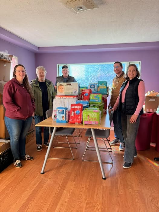 Inside a room with purple walls, a large window on the back wall, a large rectangular table with two chairs pushed in on the long sides. On top of the table is a variety of diapers and diaper wipes. Ferndale Kiwanis Club members, along with a Community Resource employee, are standing around the table smiling.