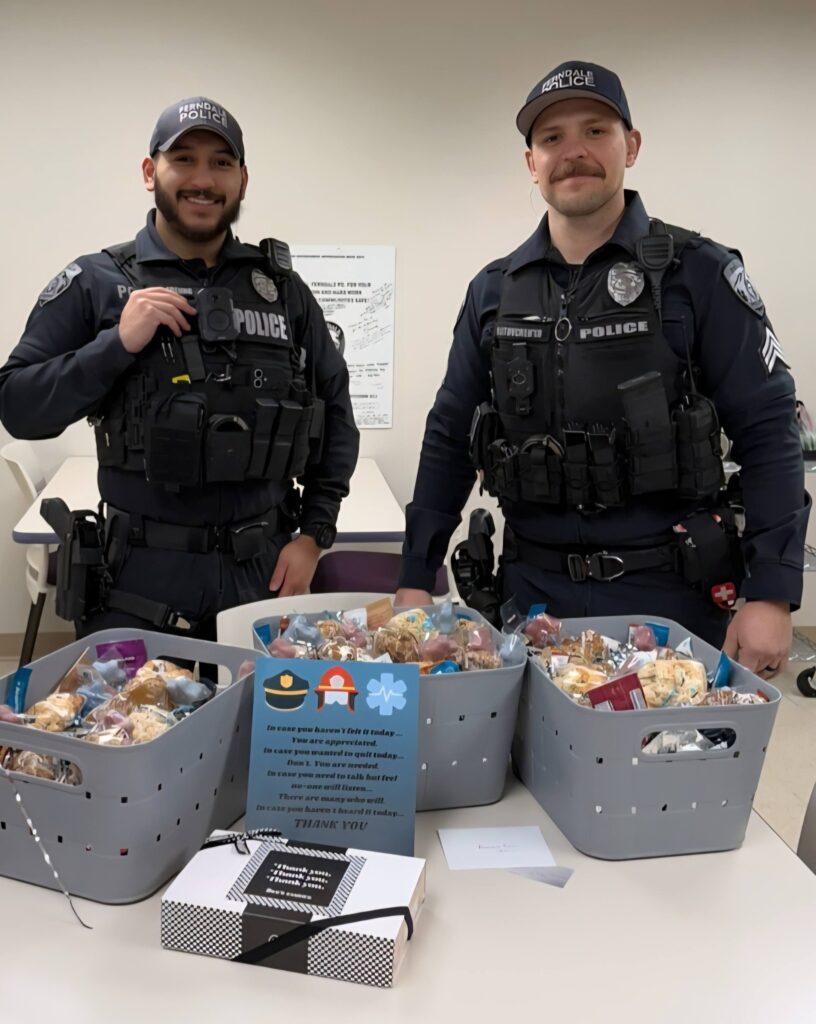 Two Ferndale police officers in uniform are standing behind a table with three baskets full of breakfast items and treats, and a box of See's peanut brittle.