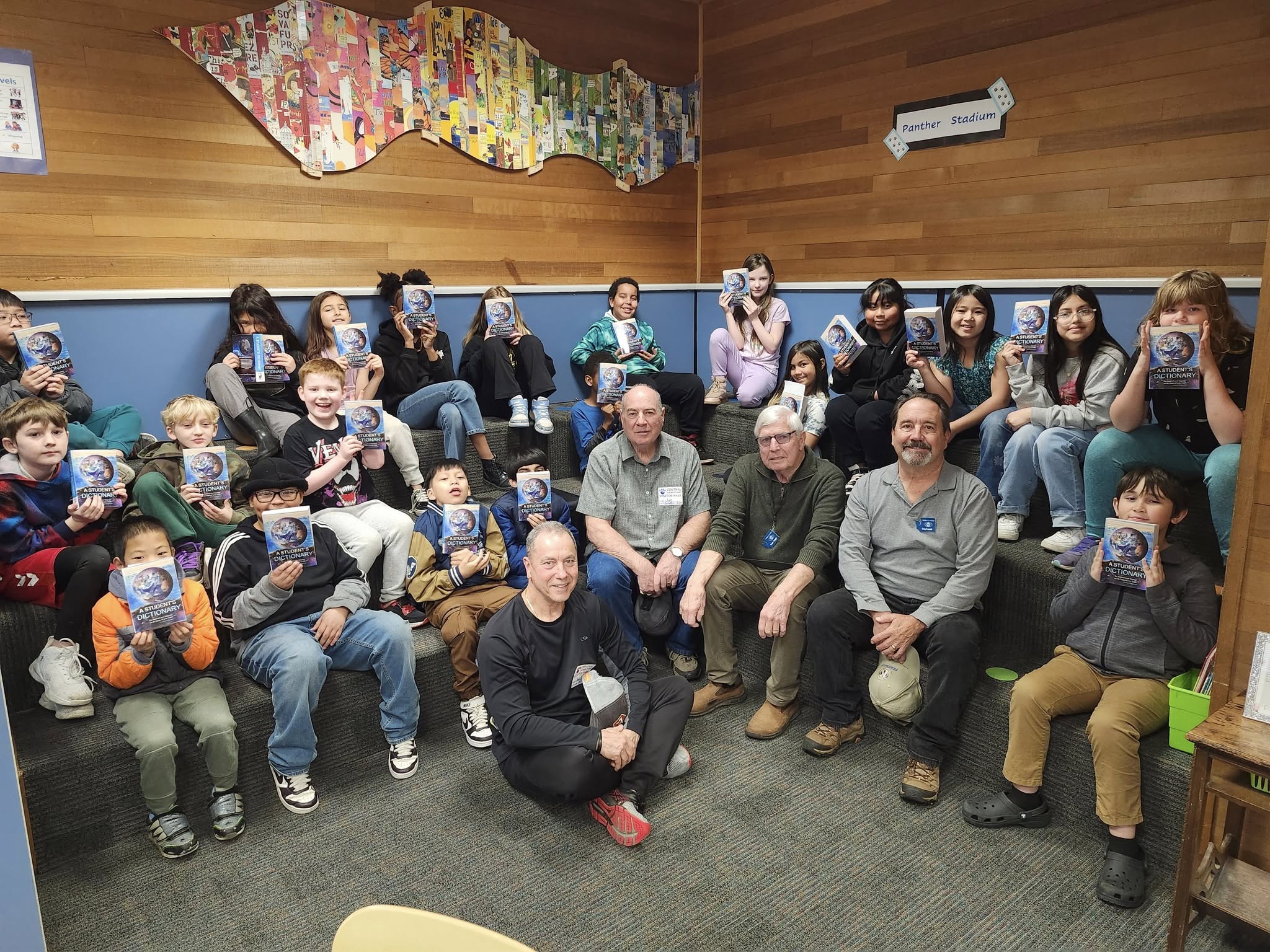 A group of third-grade students and Ferndale Kiwanis Club members are sitting on riser benches, holding up their new dictionaries.
