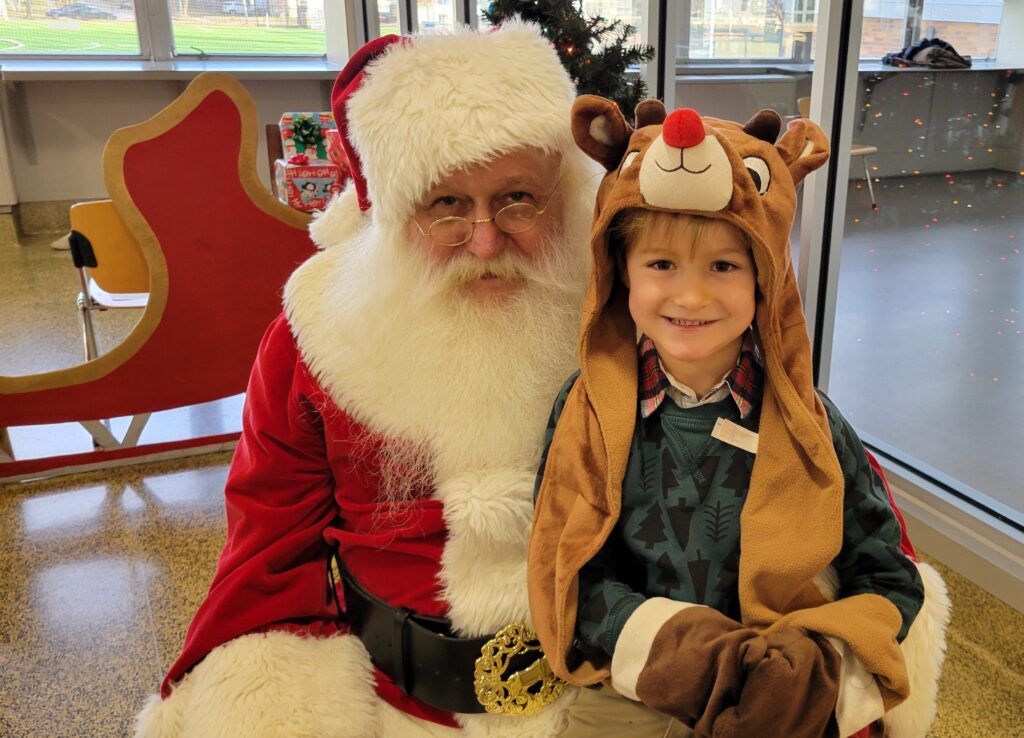 Santa with child on lap posing for picture. Child is young boy wearing a reindeer hat with long ears. 