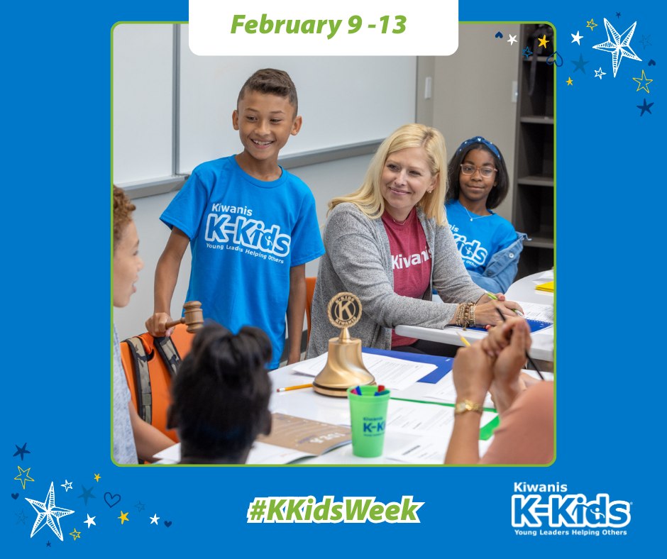 An elementary school student wearing a blue Kiwanis K-Kids shirt stands and speaks during a classroom meeting with classmates and an adult leader as part of K-Kids Week.