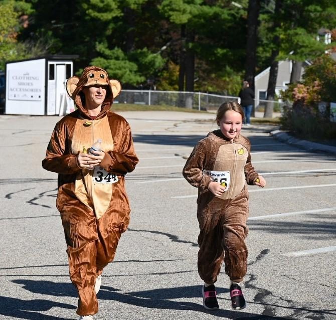 An adult and a child run across a parking lot in matching brown monkey costumes, wearing race bibs.