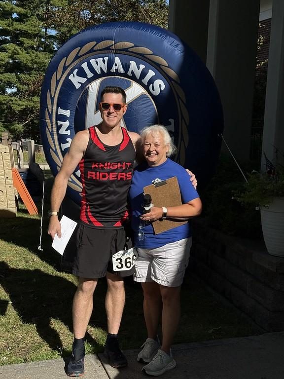 A runner wearing a race bib poses with a Kiwanis volunteer holding a clipboard in front of a large inflatable Kiwanis logo.