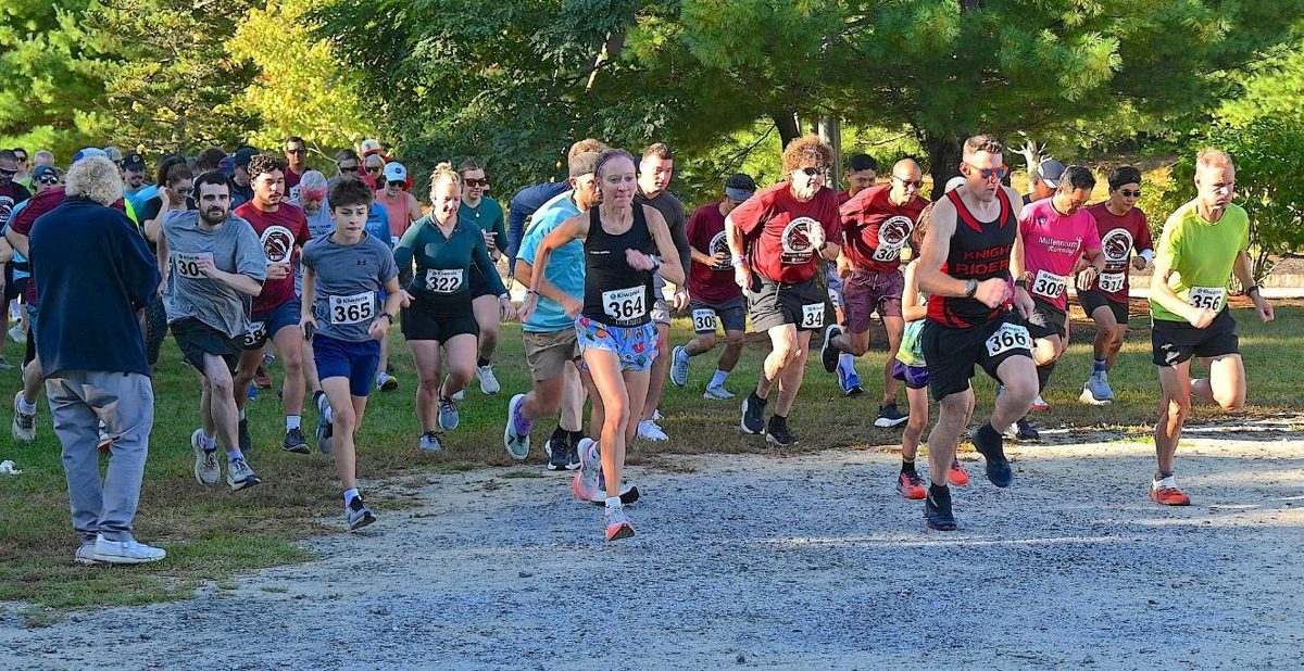 A large pack of runners wearing race bibs surges forward from the start line on a gravel path under green trees.