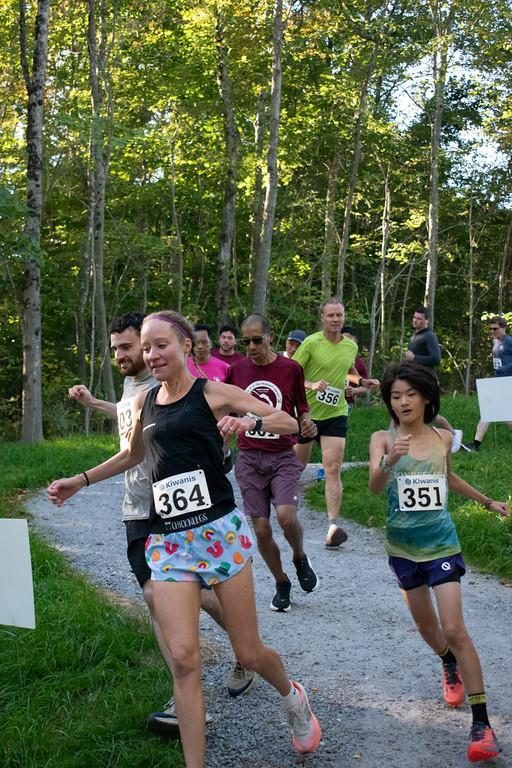 A woman and a young runner, both wearing bibs, lead a group along a gravel path lined with trees.