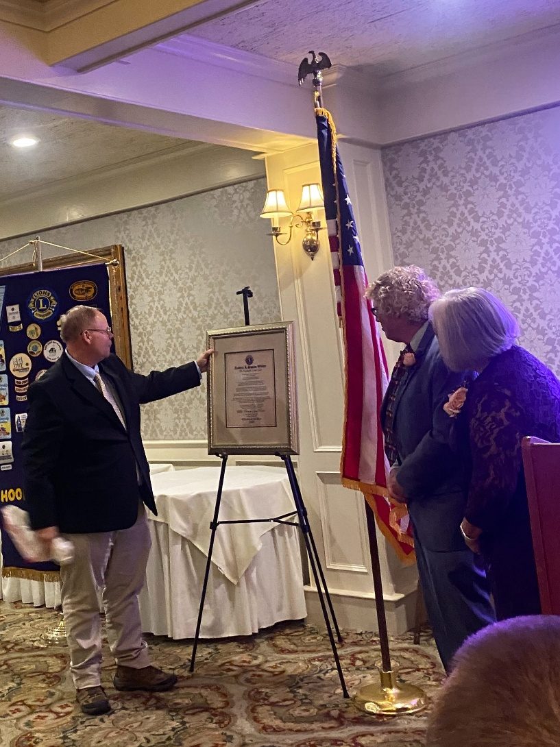 A presenter unveils a framed award on an easel for Bob and Sue Willey beside an American flag.