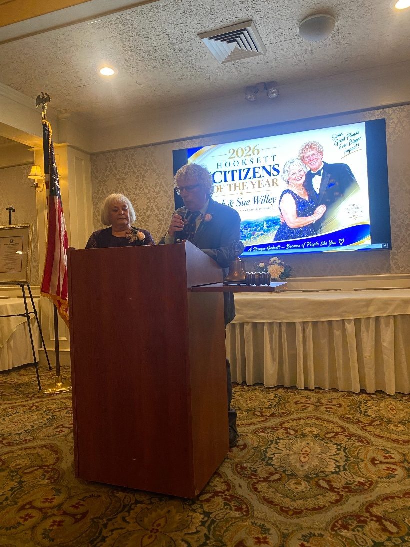 Bob Willey speaks at a podium with Sue Willey beside him, in front of a screen reading &ldquo;2026 Hooksett Citizens of the Year.&rdquo;