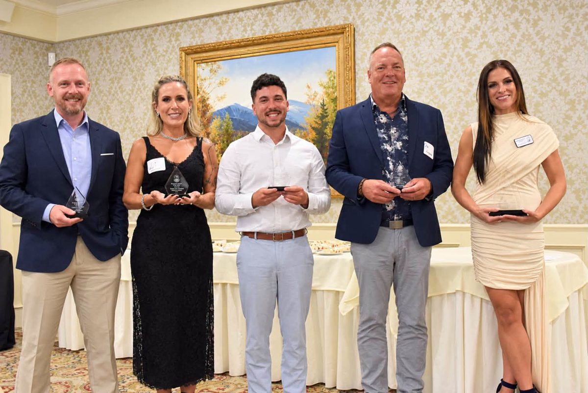 Five Chamber of Commerce award recipients in formal attire stand in a row holding their awards in a banquet room.