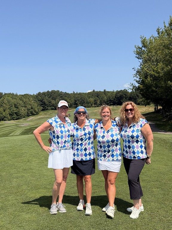 Four golfers in matching blue-and-white argyle shirts smiling together on a sunny fairway.