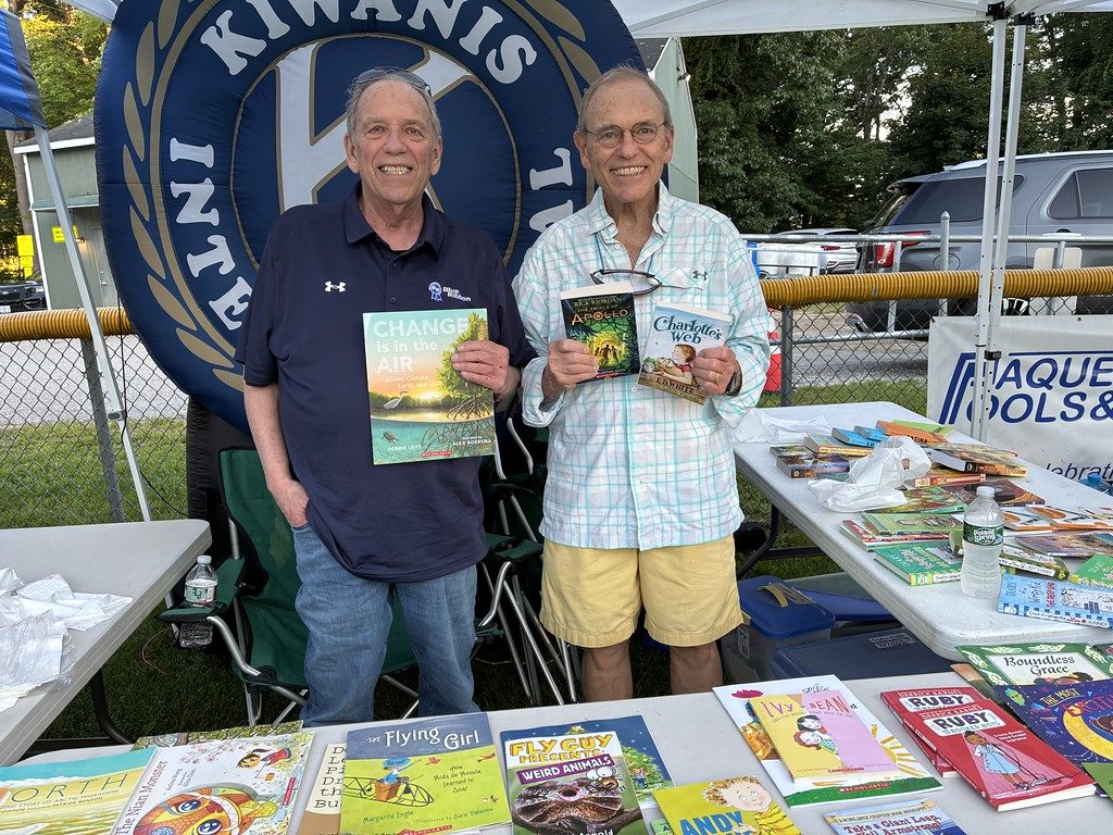 Two Kiwanis members hold up children&rsquo;s books beside a table of free books, in front of a large Kiwanis International banner.