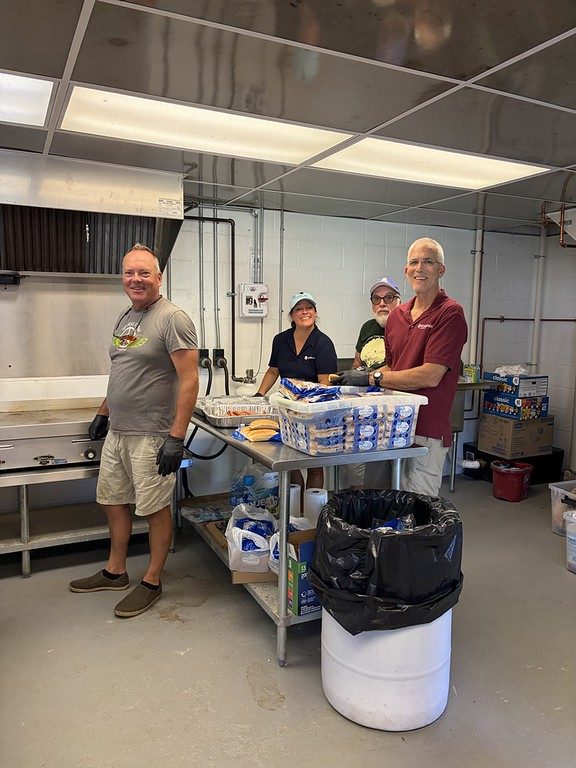 Kiwanis volunteers in a commercial kitchen preparing food at a stainless-steel prep table stacked with buns.