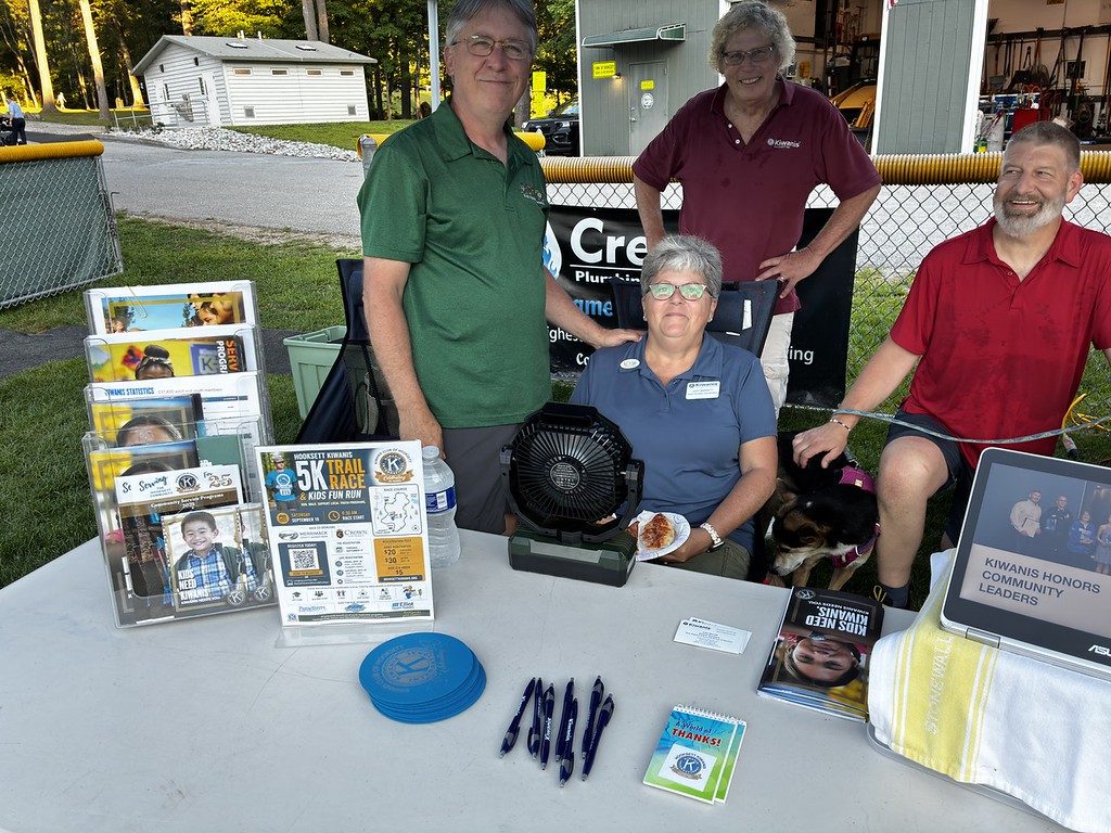 Kiwanis members seated at an outdoor information table displaying club brochures and flyers.