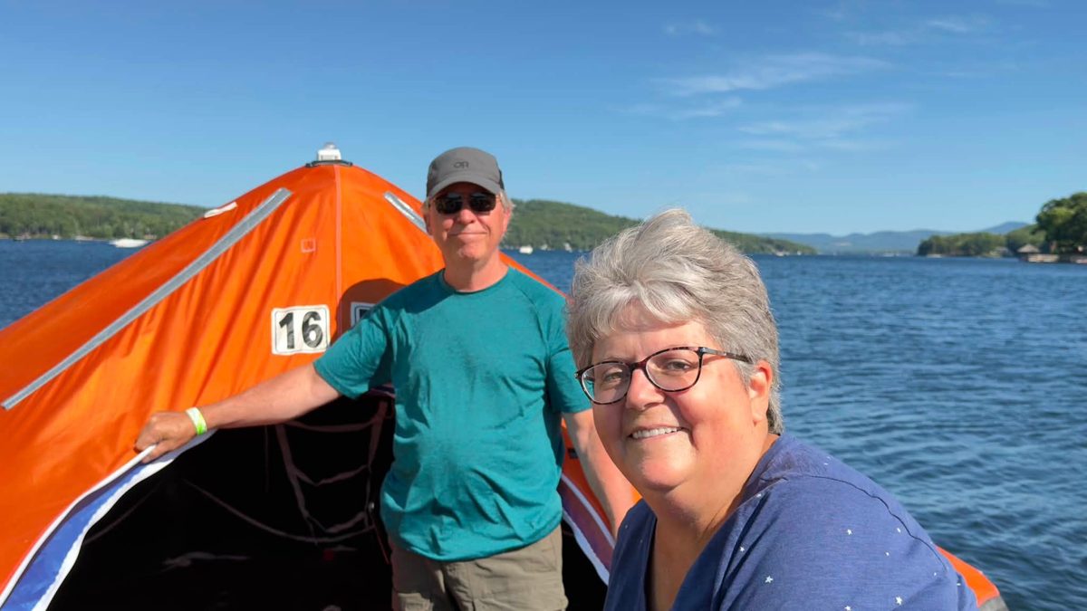 A smiling man and woman aboard an orange inflatable raft numbered 16 on Lake Winnipesaukee, with blue water and wooded hills behind them.