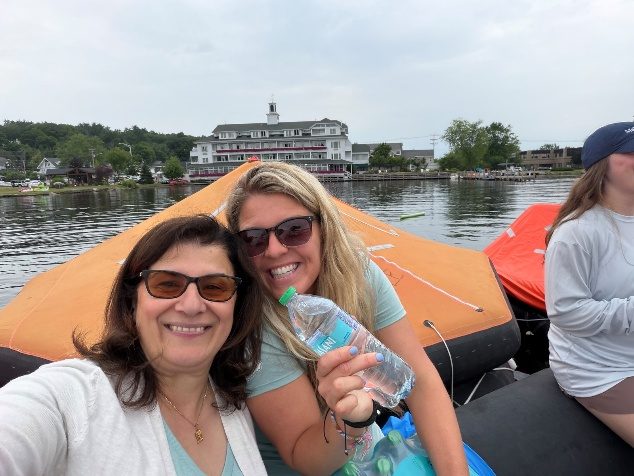 Two smiling women take a selfie on an orange raft, with Meredith&rsquo;s waterfront behind them.