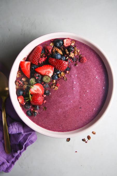 A berry smoothie in a white bowl with an assortment of berries and granola topping half of the smoothie. A purple cloth napkin with a gold spoon are beside the bowl.