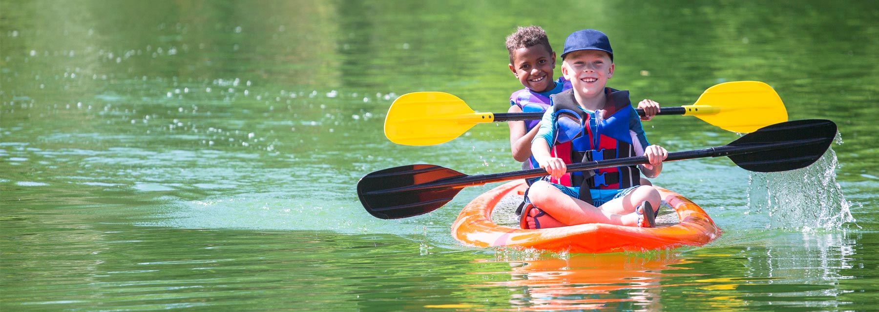 Two young smiling boys wearing multi-colored life vests while paddling in a orange kayak on a calm green lake