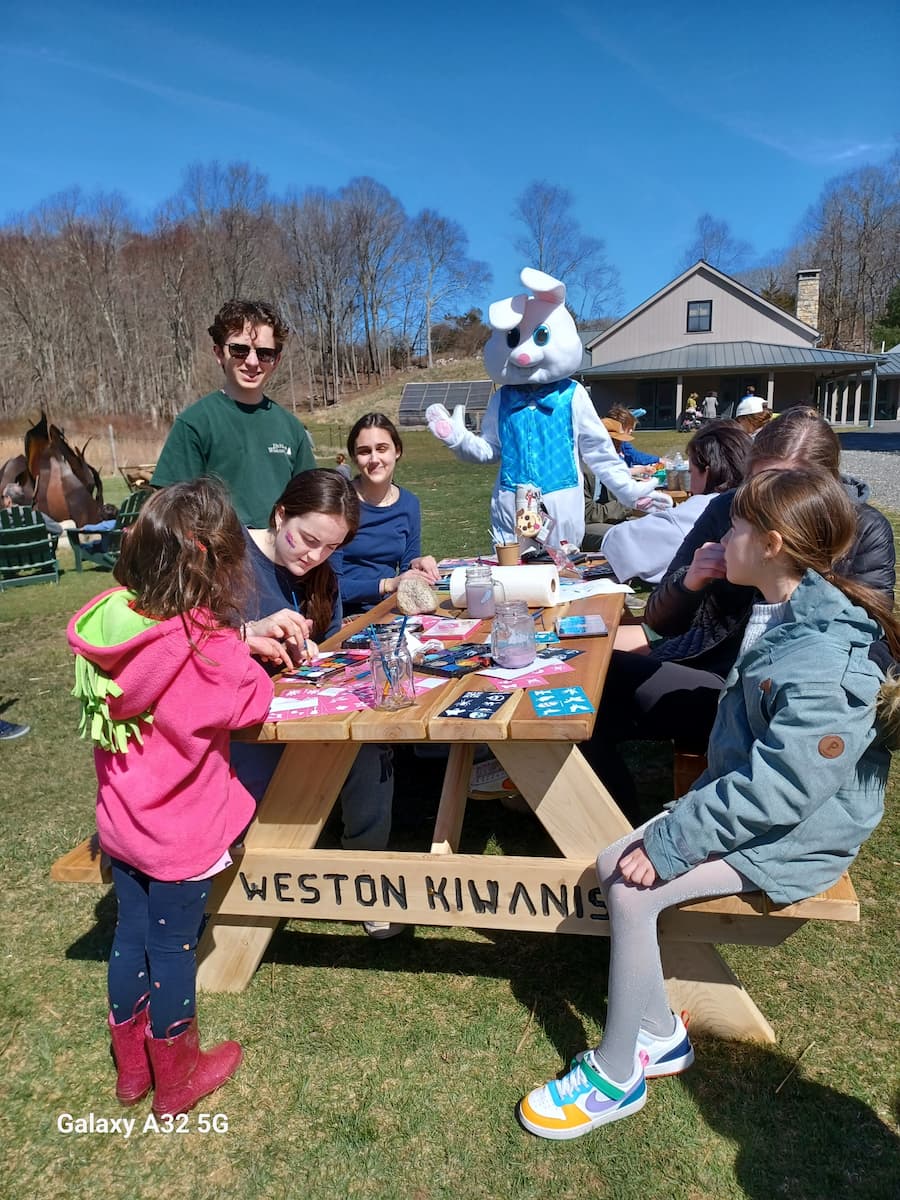 Outdoors at Lachat Town Farm on a sunny day, a family with children is engaged in crafts and is seated at a picnic table with "Weston Kiwanis" routed in the seat support. An adult costumed as a bunny is at the table.