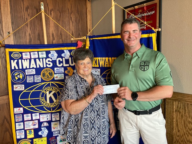 A man and woman stand in front of the club flags. The woman is handing the man a check.