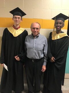 K-W Kiwanis Bursary Committee chair poses with two student scholarship awardees in their graduation robes