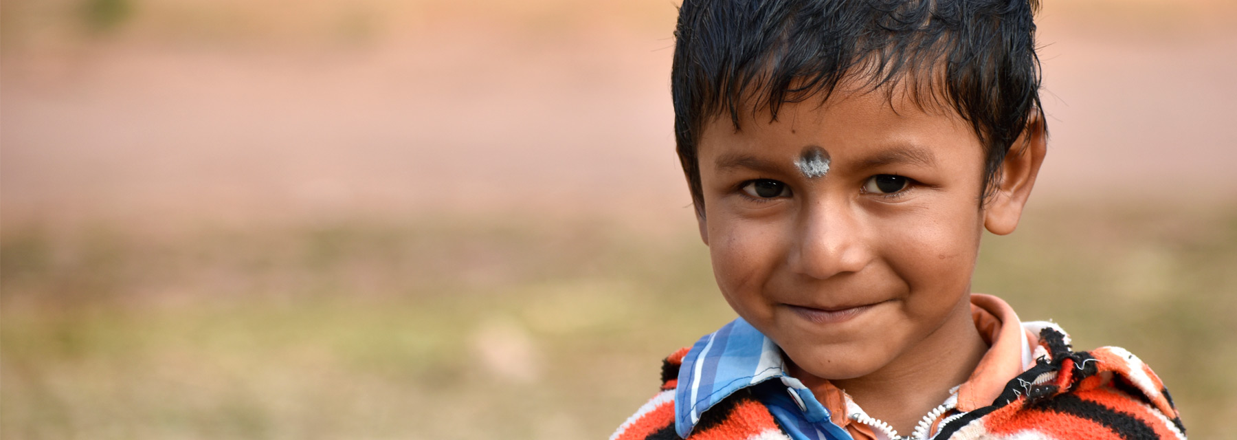 Young smiling boy with short black hair wearing a orange and brown patterned poncho in a field