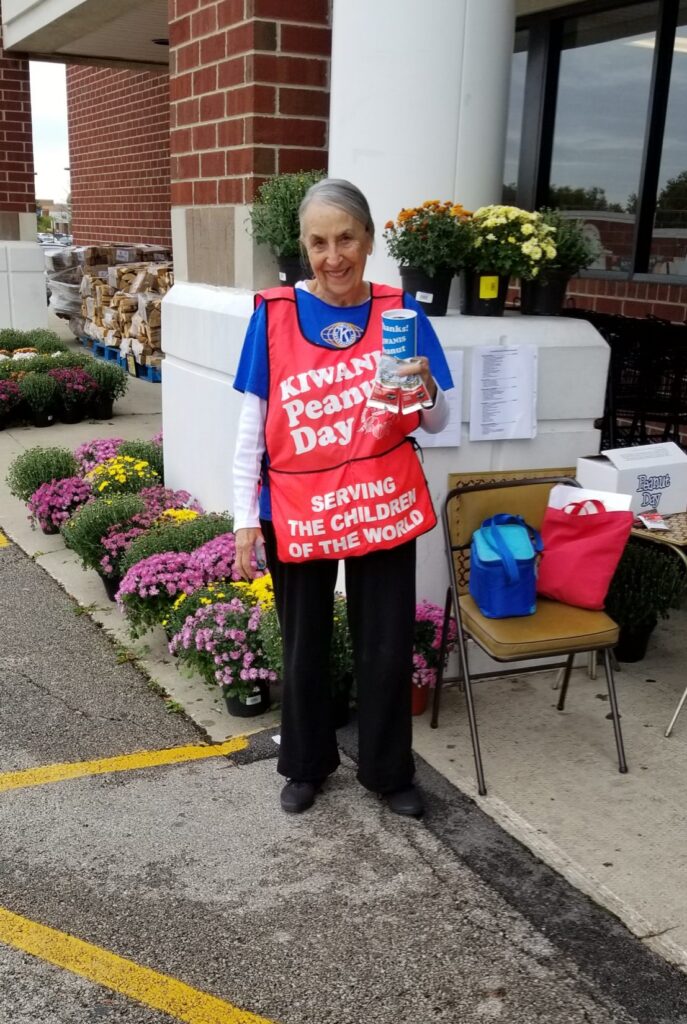 Lady selling peanuts at a store, wearing Kiwanis apron, holding donation can and bags of peanuts